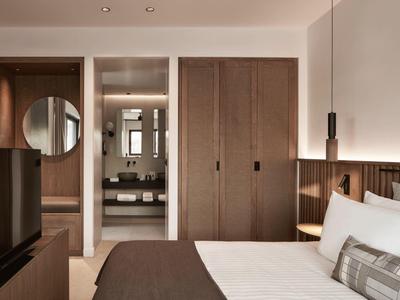 Modernes Schlafzimmer in gedeckten Farben mit Holzmöbeln und Blick ins Bad mit Waschbecken und Spiegel.