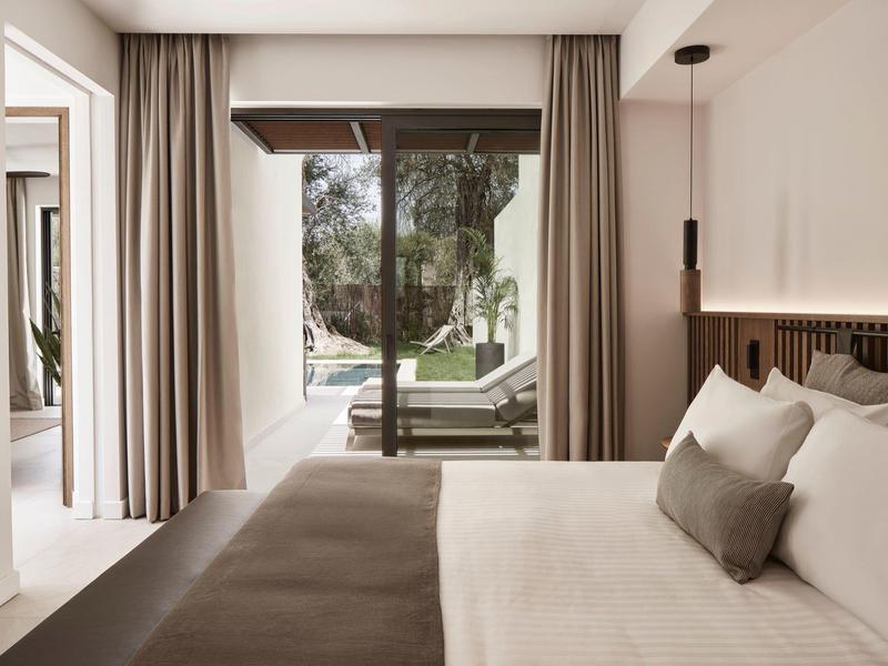 Světý pokoj hotelu s manželskou postelí, velkými okny a výhledem do zahrady s lehátky.