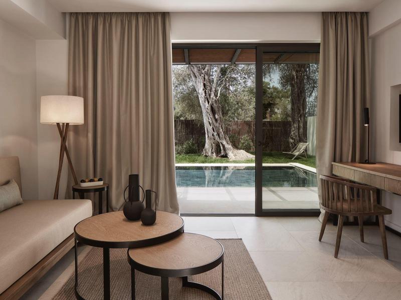Moderní hotelový obývací pokoj s béžovou pohovkou, kulatými stolky, psacím stolem a výhledem na bazén.