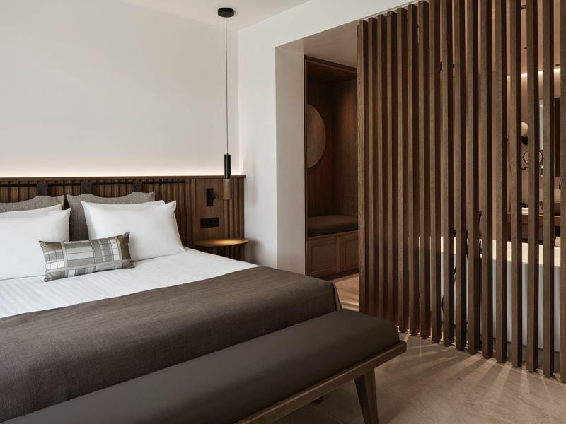 Moderní hotelový pokoj s manželskou postelí, dřevěnou lavicí a dřevěnou příčkou oddělující koupelnu.