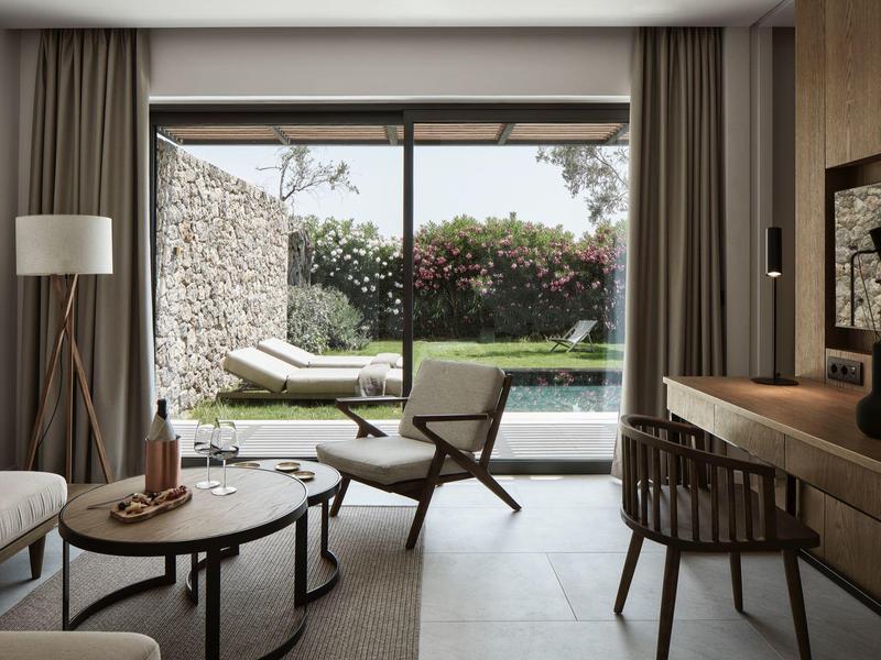 Světý obývací pokoj se sedacími křesly, stolkem, psacím stolem a výhledem na zahradu a bazén.