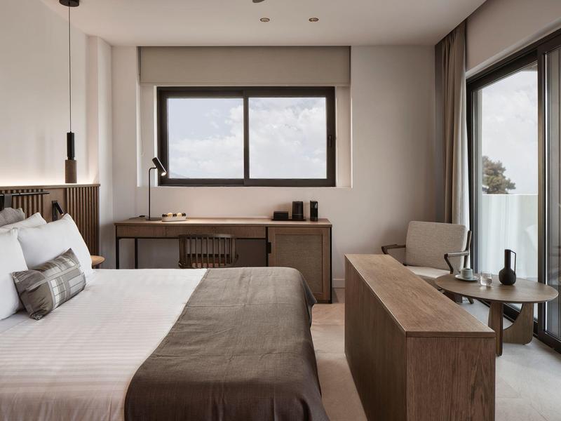 Moderní hotelový pokoj s manželskou postelí, psacím stolem, křeslem a velkým oknem.