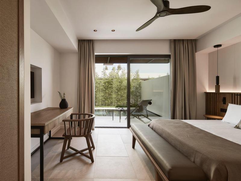 Moderní hotelový pokoj s manželskou postelí, psacím stolem, dřevěnou židlí a výhledem na balkon a zahradu.