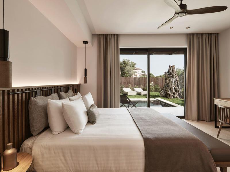 Moderní hotelový pokoj s manželskou postelí, pracovním stolem, stropním ventilátorem a výhledem na zahradu.