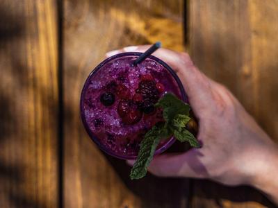 Mano che tiene un bicchiere di frullato viola guarnito con menta e cannucce su superficie di legno.