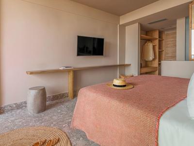 Camera d'hotel semplice con copriletto rosa, TV a parete e tavolo in legno minimalista.