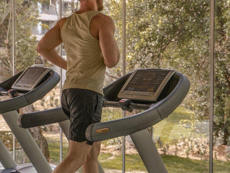 Uomo che si allena su un tapis roulant in una palestra con grandi finestre che si affacciano sul giardino.