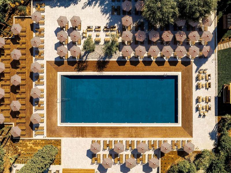 Vista aerea di una piscina rettangolare con lettini e ombrelloni, circondata da alberi e verde.