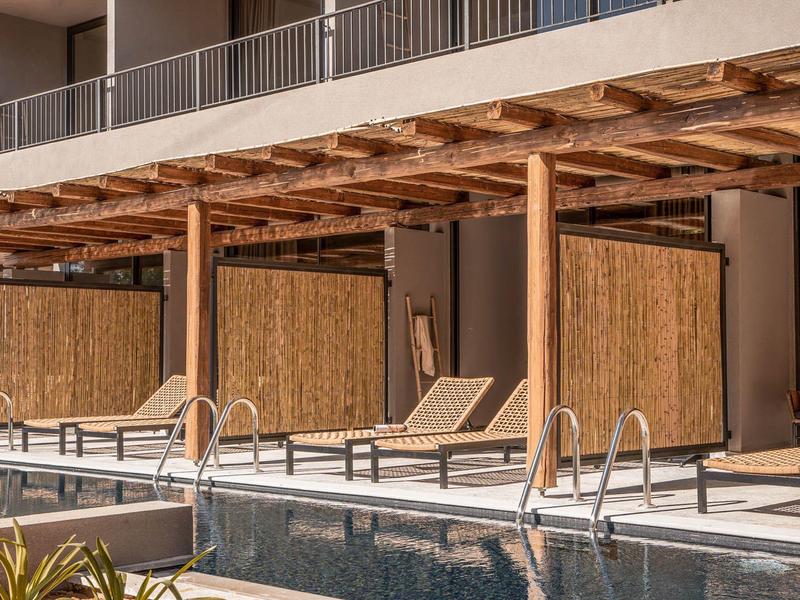 Pergolato in legno con lettini accanto a una piscina davanti a un hotel.