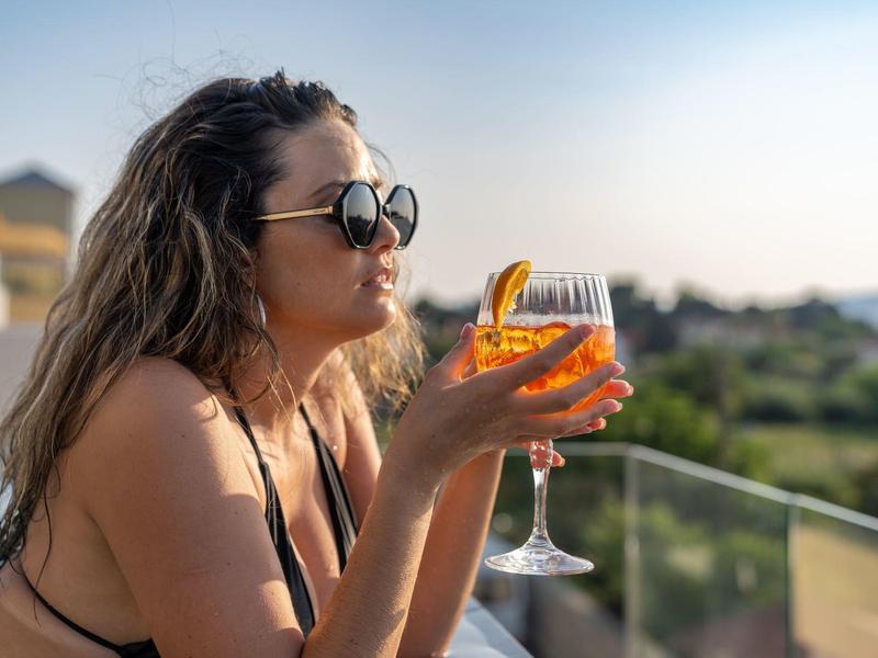 Donna con occhiali da sole che tiene un bicchiere di vino su una terrazza dell'hotel al tramonto.
