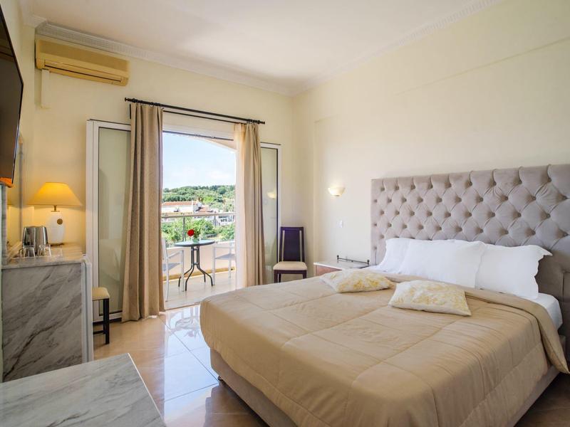 Světle zařízený hotelový pokoj s postelí, balkonem a výhledem na zelenou krajinu.