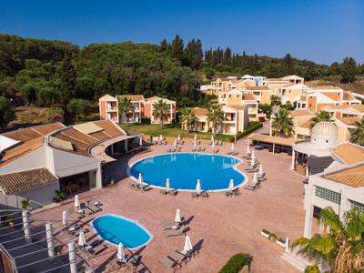 Hotelanlage mit rundem und ovalem Pool, Sonnenliegen und Gebäuden vor bewaldetem Hang bei klarem Himmel.