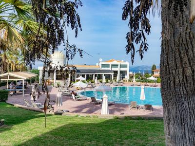 Ein Hotel mit Außenpool, umgeben von Liegestühlen, Palmen und grünem Rasen bei blauem Himmel.