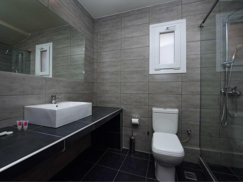 Modernes Badezimmer mit grauen Fliesen, großer Spiegel, weißem Waschbecken, WC und Glasdusche.