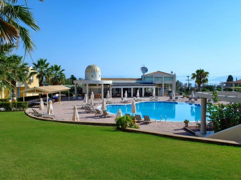 Großer Pool mit blauer Wasserfläche, umgeben von Liegen, Palmen und grünem Rasen bei klarem Himmel.