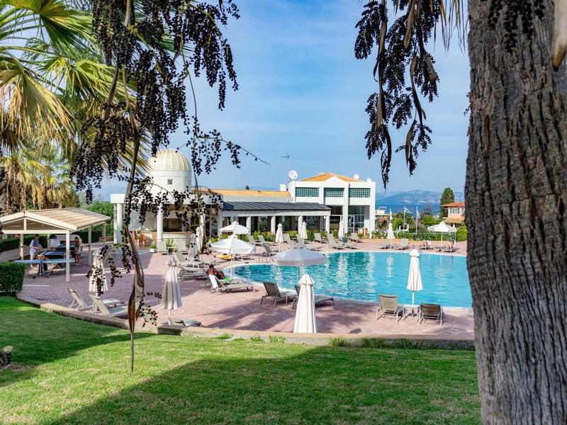 Poolbereich mit Liegestühlen, Sonnenschirmen und Palmen vor einem weißen Hotelgebäude bei blauem Himmel.