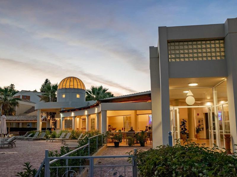 Moderner Gebäudekomplex mit Glaskuppel, Außenbeleuchtung und bewaldeter Umgebung bei Dämmerung.