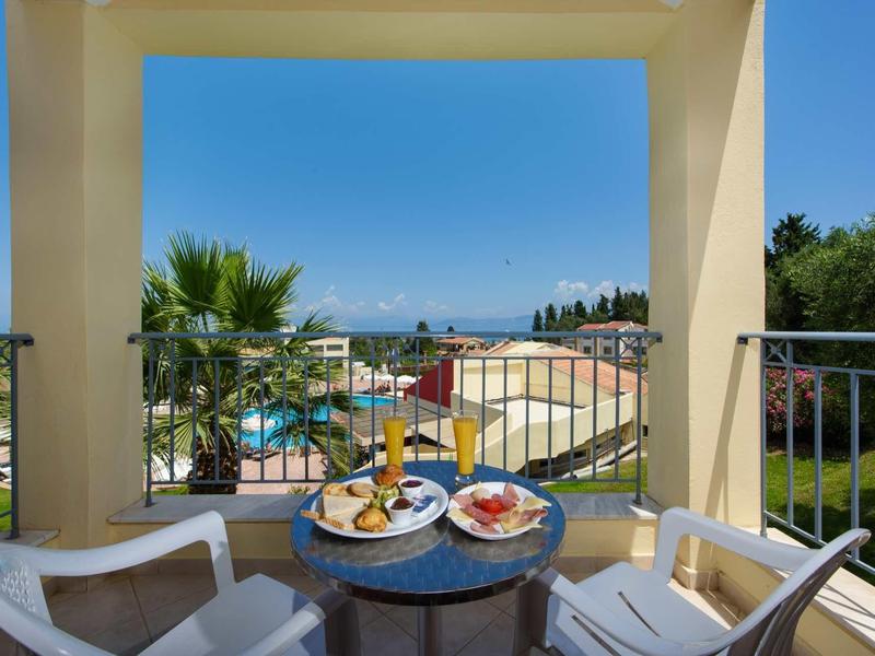 Balkon mit Frühstückstisch und zwei Stühlen, Blick auf Pool, Palmen und blauen Himmel.
