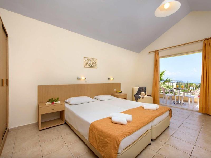 Helles Schlafzimmer mit Doppelbett, beigen Wänden, Fliesenboden und Balkon mit Meerblick.