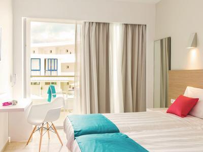 Bright hotel room with double bed, white furniture, and balcony overlooking the sea.