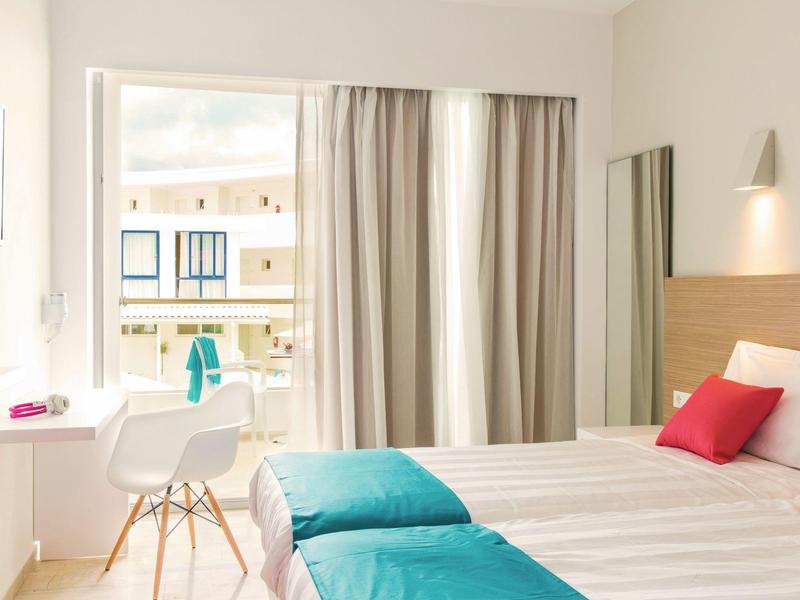 Bright hotel room with double bed, white furniture, and balcony overlooking the sea.