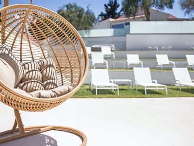 Moderner hängender Korbsessel mit Kissen vor einer Liegewiese mit weißen Liegestühlen.