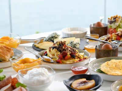 Een ontbijt tafel gedekt met omelet, broodjes, fruit en diverse bijgerechten bij een raam met uitzicht op water.