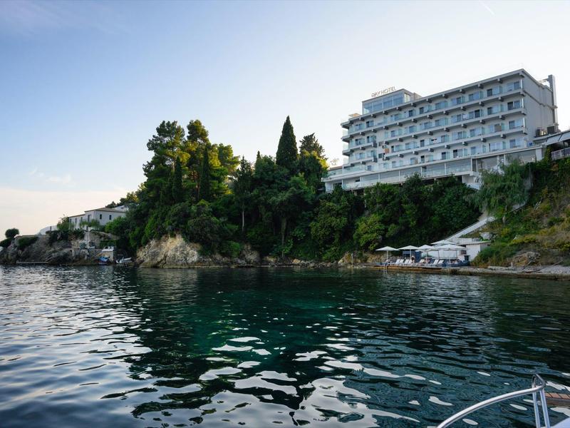 Groot hotelgebouw aan rotsachtige kust met kalme zee op de voorgrond onder een heldere avondlucht.