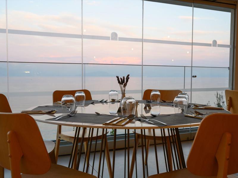 Elegant restaurant met panoramisch zeezicht door grote ramen bij zonsondergang.