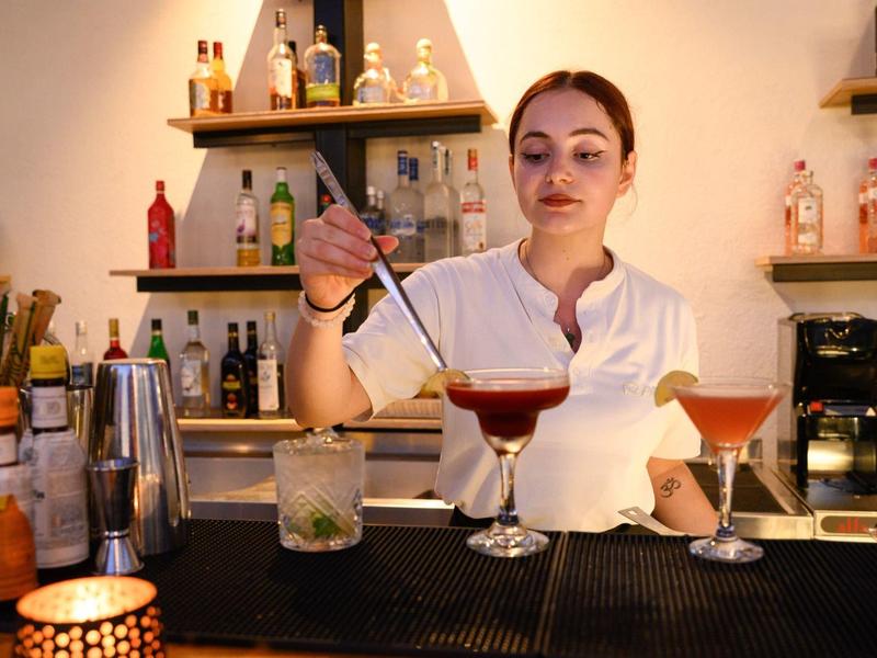 Een barman bereidt twee kleurrijke cocktails voor bij een bar met diverse flessen op de achtergrond.
