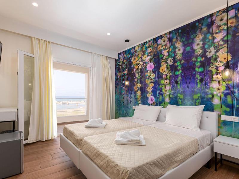 Moderní hotelový pokoj s manželskou postelí, květinovým uměním na stěně a výhledem na moře oknem.