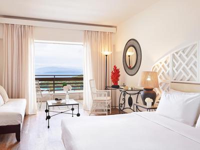 Chambre d'hôtel lumineuse avec grand lit, fauteuils et balcon avec vue sur la mer.