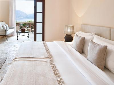 Slaapkamer met groot bed, kussens, tafellamp en uitzicht op terras en zee.