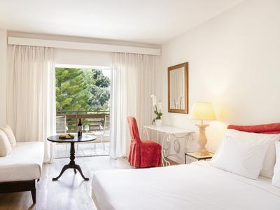 Chambre d'hôtel lumineuse avec lit double, bureau, chaise rouge et balcon avec vue sur les arbres.