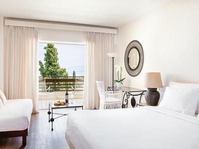 Chambre d'hôtel lumineuse avec lit double, canapé, bureau et balcon avec vue sur des arbres verts.