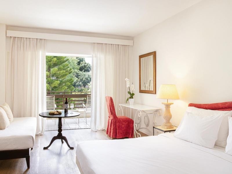 Chambre d'hôtel lumineuse avec lit double, bureau, chaise rouge et balcon avec vue sur les arbres.