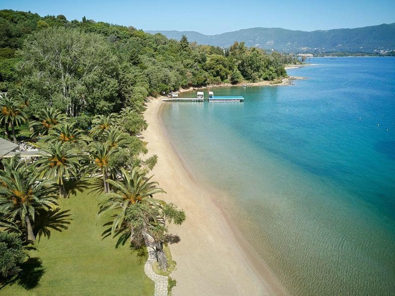 Plage de sable avec eau claire, entourée de palmiers et de collines boisées sous un ciel bleu.