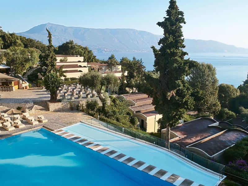 Vue d'une piscine d'hôtel avec des chaises longues, des arbres et la mer en arrière-plan par temps clair.