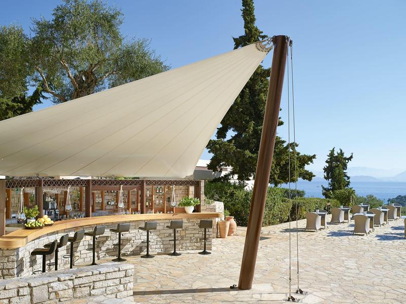 Terrasse avec grande voile d'ombrage, tables et chaises à côté d'un bâtiment en pierre avec vue sur la mer.