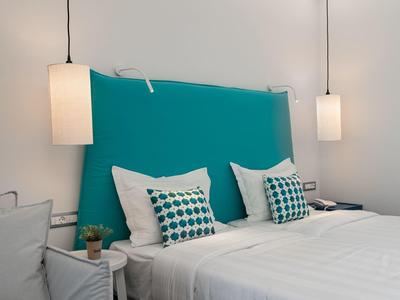Weißes Bett mit türkisfarbenem Kopfteil, zwei gemusterten Kissen und Hängelampen an weißer Wand.