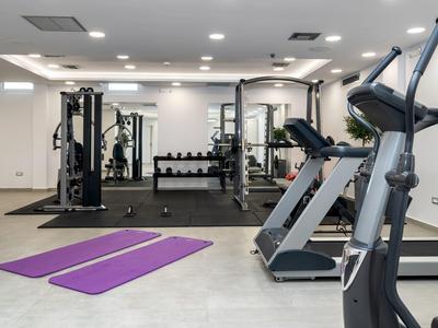 Moderner Fitnessraum mit Trainingsgeräten, Hanteln, Spiegeln und zwei lila Yogamatten auf grauem Boden.