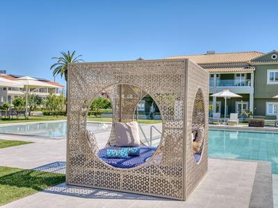 Moderner Lounge-Würfel mit Kissen neben einem Hotelpool und Grünflächen unter blauem Himmel.