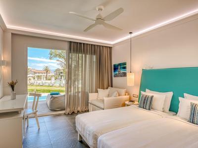 Helles Hotelzimmer mit Doppelbett, Sofa, Schreibtisch und Balkon mit Blick auf Garten und Pool.