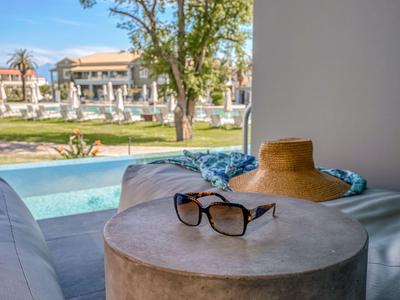Sonnenbrille und Hut auf einem runden Beistelltisch neben einem Pool mit Blick auf Hotel und Garten.