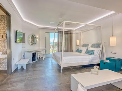 Helles Hotelzimmer mit Himmelbett, Sitzecke, TV und Balkon mit Meerblick.