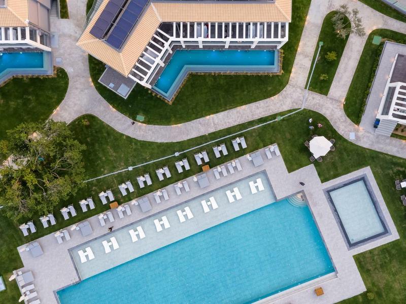 Letecký pohled na hotelový bazén s lehátky a slunečníky na trávníku.