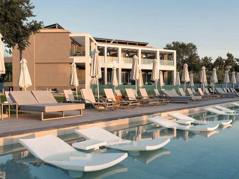 Moderní hotelový bazén s lehátky a slunečníky před bílou budovou za jasné oblohy.