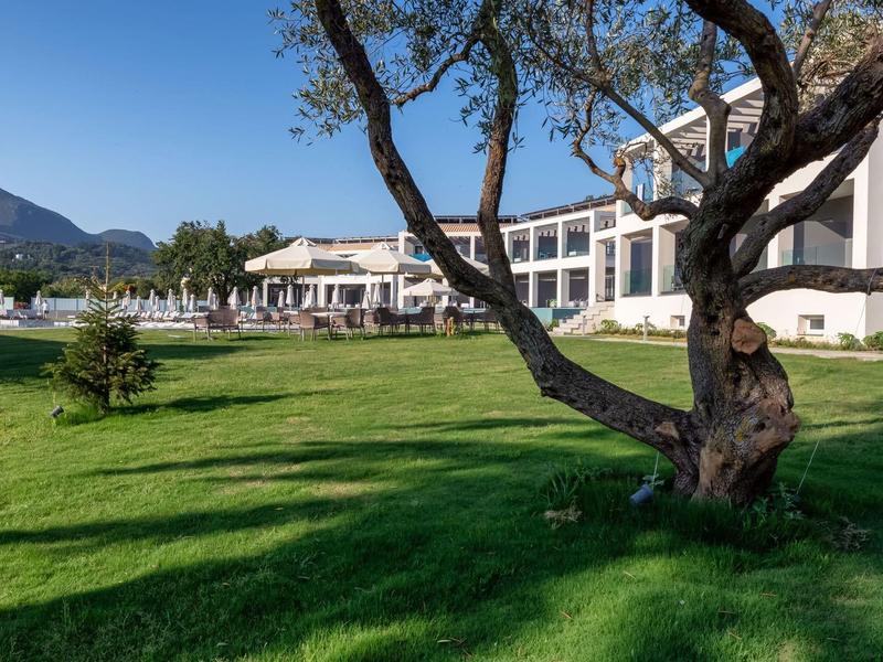 Velká budova hotelu s terasou, obklopená zeleným trávníkem a stromy pod jasnou oblohou.