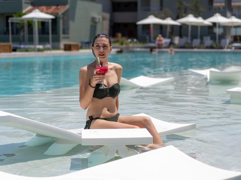 Žena v černých bikinách sedí u bazénu na lehátku a drží červený nápoj.