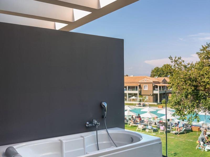 Volně stojící vana na balkóně s výhledem na hotelový bazén a zahradu za jasné oblohy.